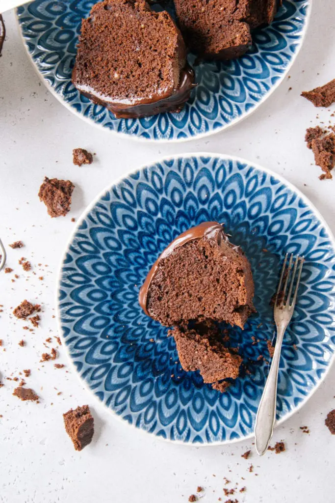 Ein Stück saftiger Schokoladenkuchen von oben auf einem blauen Teller fotografiert. Ein kleiner Teil wurde mit einer alten Kuchengabel abgestochen. Kuchenbrösel auf weißem Untergrund als Verzierung.