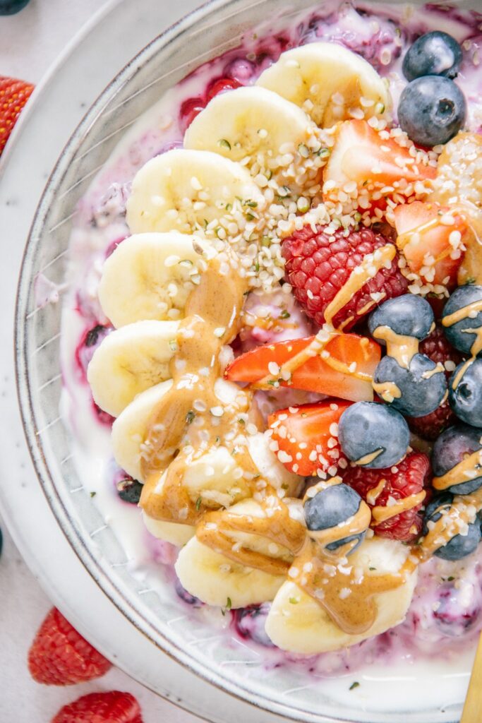 Nahaufnahme von Beeren-Bananen-Müsli von oben. Fächerartig aufgelegte Bananenscheiben mit viel Mandelmus und Beeren als Topping.