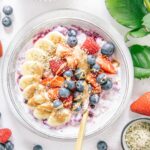 Schnelles Beeren-Bananen-Müsli mit Topping in einer graublauen Schüssel von oben fotografiert. Beeren und eine weiße Rose als Dekoration.