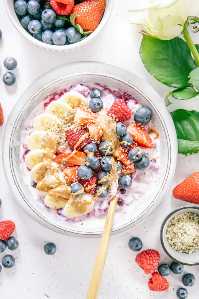 Schnelles Beeren-Bananen-Müsli mit Topping in einer graublauen Schüssel von oben fotografiert. Beeren und eine weiße Rose als Dekoration.