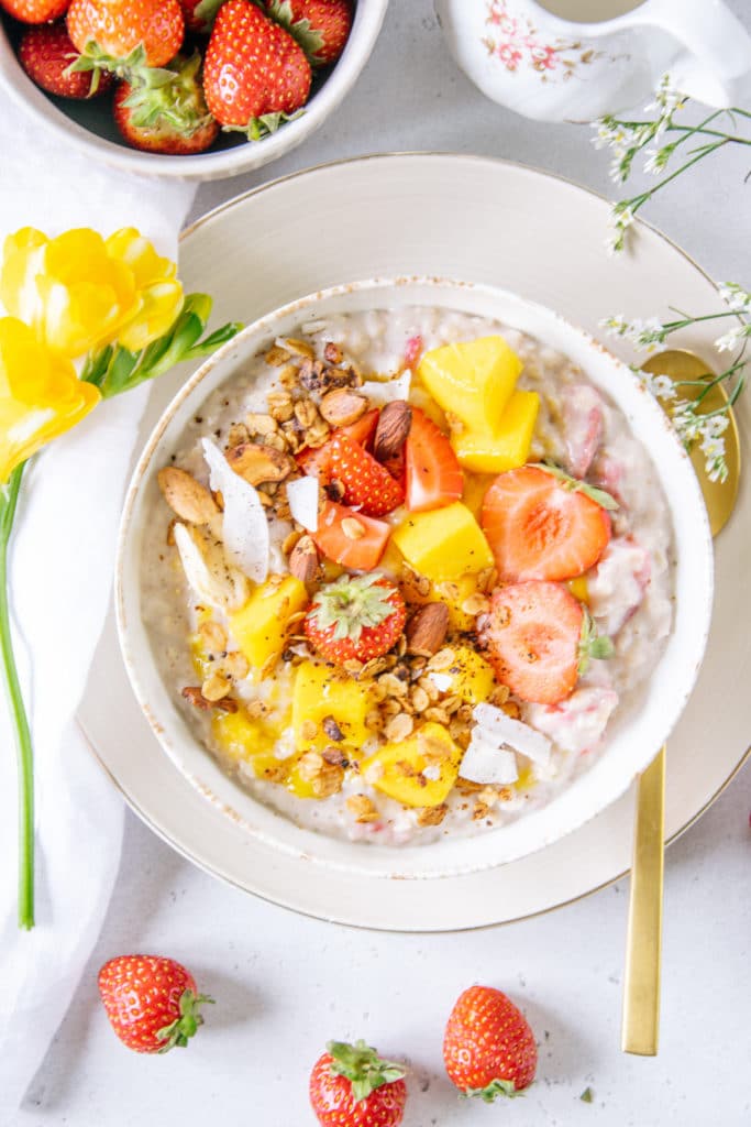 Erdbeer-Mango-Porridge in einer weißen Schüssel. Dekoriert mit Erdbeeren, Mangowürfeln und einer gelben Blume.