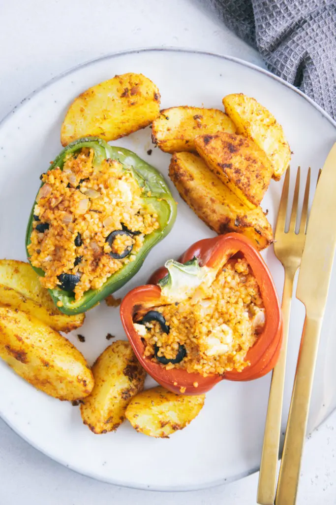 Gefüllte Bulgur-Feta-Paprika mit Ofen-Wedges auf einem graublauen Teller mit goldenem Besteck von oben fotografiert.