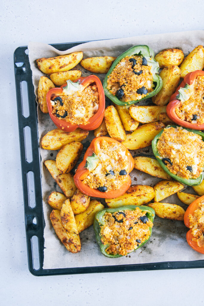 Gefüllte Bulgur-Feta-Paprika mit Ofen-Wedges auf dem Backblech von oben auf hellem Untergrund fotografiert. Blech auf der rechten Seite angeschnitten.