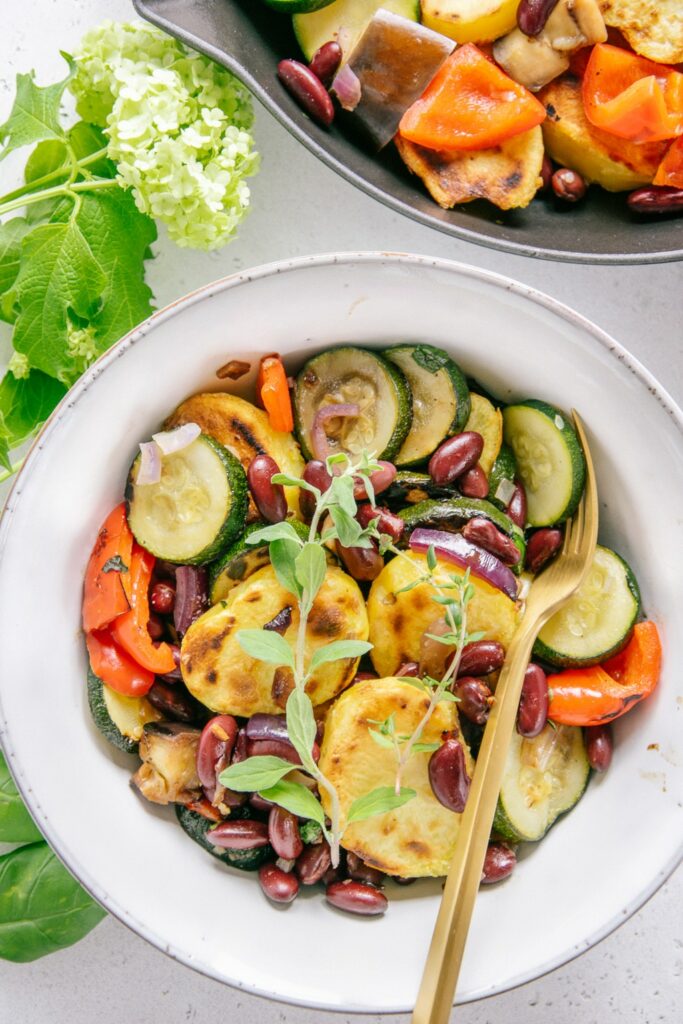 Mediterrane Bratkartoffel-Gemüsepfanne in einer graublauen Bowl mit goldenem Löffel von oben fotografiert. Dekoriert mit fischem Oregano.