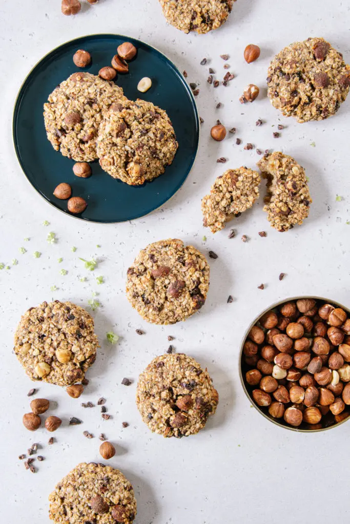 Mehrere gesunde vegane Guten Morgen Cookies verteilt auf weißem Untergrund, dekoriert mit Haselnüssen und Kakaonibs
