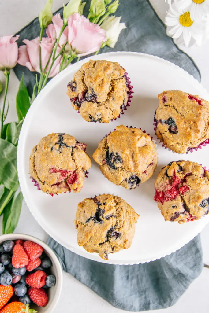 Vegane zuckerfreie Beerenmuffins auf einer weißen Tortenplatte von oben fotografiert. Eine Schüssel Beeren als Dekoration.