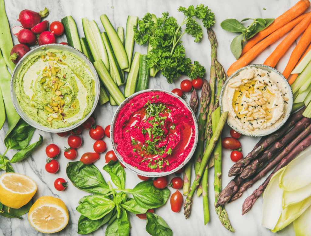 Avocado-Hummus, Rote-Bete-Hummus und klassischer Hummus in drei Schüsseln mit Rohkost drum herum auf hellem Untergrund aufgenommen