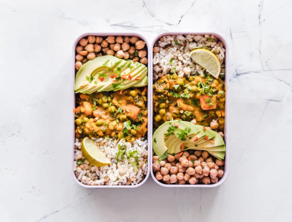 Zwei längliche eckige Lunchboxen mit Kichererbsen, Avocadospalten, Curry und Reis aus der Vogelperspektive auf hellem Untergrund aufgenommen