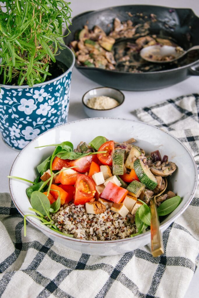 Cremige vegane Pilzpfanne mit Zucchini, Räuchertofu, Tomaten und Quinoa in einer graublauen Schüssel auf einem karierten Küchentuch. Aufnahme im 45-Grad-Winkel. Blumentopf mit Thymian als Dekoration.