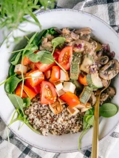Nahaufnahme von cremiger veganer Pilzpfanne mit Quinoa, Räuchertofu, Tomaten und Jungspinat in einer graublauen Bowl mit goldenem Löffel.