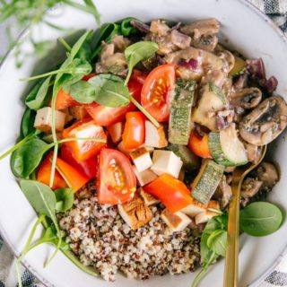 Nahaufnahme von cremiger veganer Pilzpfanne mit Quinoa, Räuchertofu, Tomaten und Jungspinat in einer graublauen Bowl mit goldenem Löffel.