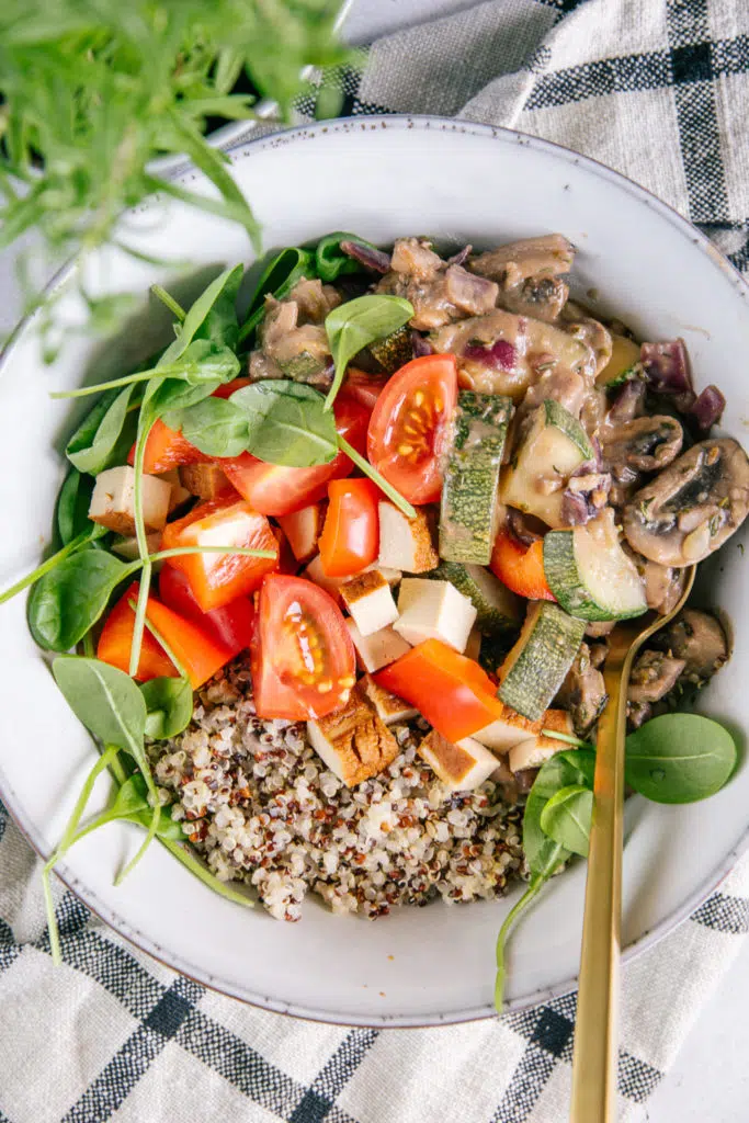 Nahaufnahme von cremiger veganer Pilzpfanne mit Quinoa, Räuchertofu, Tomaten und Jungspinat in einer graublauen Bowl mit goldenem Löffel.