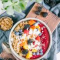 Porridge mit Nicecream in einer graublauen Schüssel auf einem dunklen Holzbrett und einem blauen Leinentuch. Dekoration aus Heidelbeeren, Brombeeren, Dinkelflakes und einer Grünpflanze.