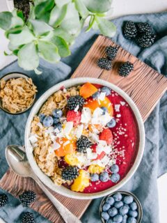 Porridge mit Nicecream in einer graublauen Schüssel auf einem dunklen Holzbrett und einem blauen Leinentuch. Dekoration aus Heidelbeeren, Brombeeren, Dinkelflakes und einer Grünpflanze.