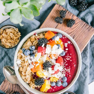 Porridge mit Nicecream in einer graublauen Schüssel auf einem dunklen Holzbrett und einem blauen Leinentuch. Dekoration aus Heidelbeeren, Brombeeren, Dinkelflakes und einer Grünpflanze.