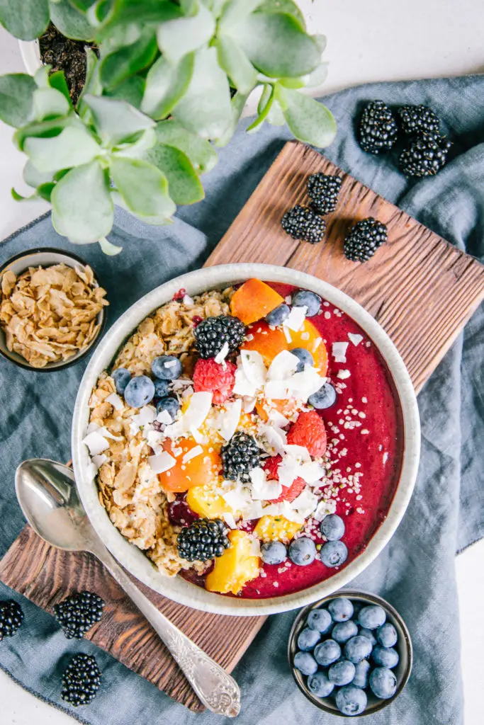 Porridge mit Nicecream in einer graublauen Schüssel auf einem dunklen Holzbrett und einem blauen Leinentuch. Dekoration aus Heidelbeeren, Brombeeren, Dinkelflakes und einer Grünpflanze.