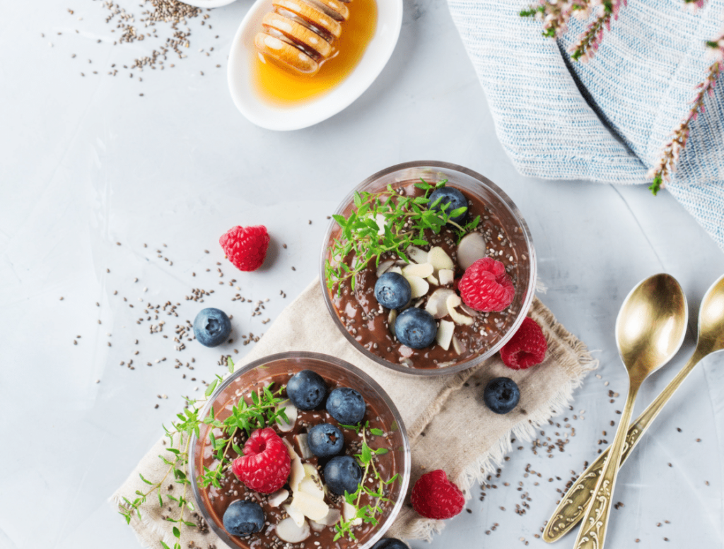 Veganer Schokoladenpudding in zwei Gläsern garniert mit Beeren, Mandelblättchen und frischem Thymian auf einem braunen Tuch mit goldenen Löffeln. Aufnahme aus der Vogelperspektive