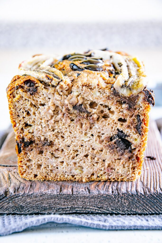 Nahaufnahme von Chocolate Chip Bananenbrot in der Frontalansicht auf einem rustikalen Holzbrett. Die saftige Teigstruktur ist deutlich erkennbar.