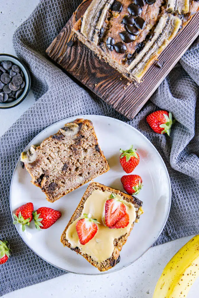 Zwei Scheiben gesundes veganes Chocolate Chip Bananenbrot auf einem graublauen Teller. Eine davon ist mit Mandelmus bestrichen und mit Erdbeeren belegt. Aufnahme von oben.