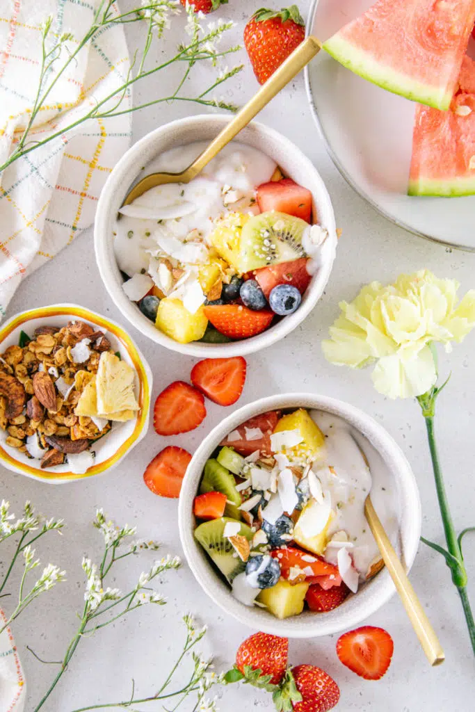 Zwei kleine Obstsalat Joghurt Bowls auf hellem Untergrund, verziert mit Erdbeeren und Granola