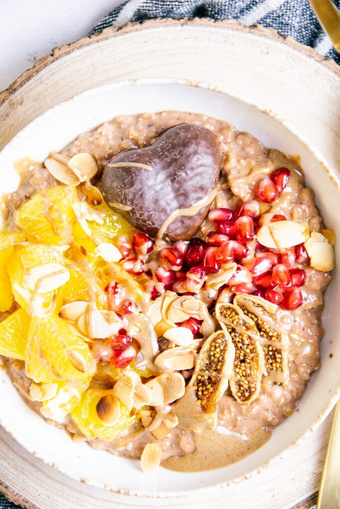 Lebkuchen-Porridge mit Orange in einer hellen Schüssel auf einem Rindenbrett. Getrocknete Feige, Granatapfelkeren, Mandelblättchen, Orangenstücke und Lebkuchen als Toppings.