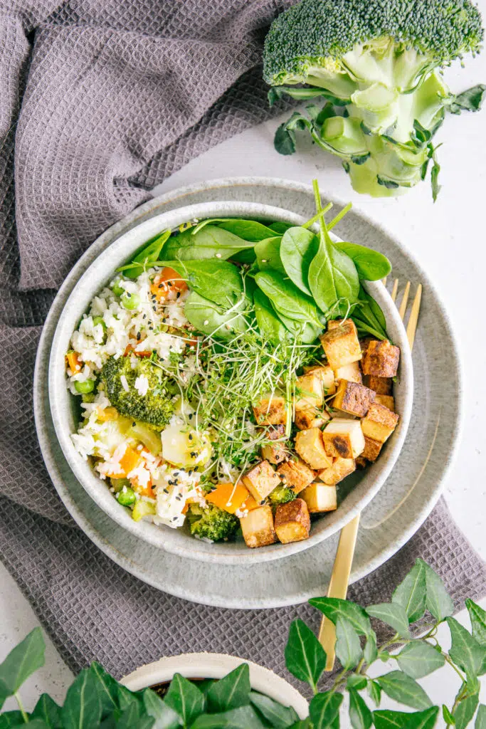 Gemüsereis mit gebratenem Tofu in einer graublauen Schüssel auf dazu passendem Teller mit einer goldenen Gabel darauf. Graues Küchentuch als Untergrund.