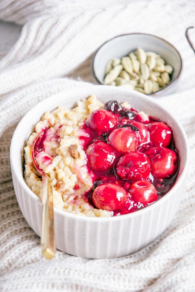 Veganer und zuckerfreier Milchreis mit Kirschkompott in einer grauen Schüssel mit goldenem Löffel im 45-Grad-Winkel fotografiert.