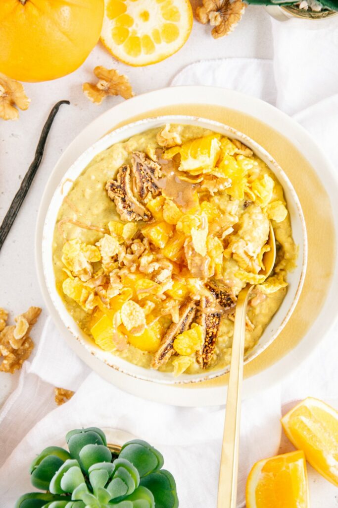 Orangen-Vanille Protein-Porridge getoppt mit Orangen, Cornflakes, Walnüssen, getrockneten Feigen und Haselnussmus. Goldener Löffel im Porridge