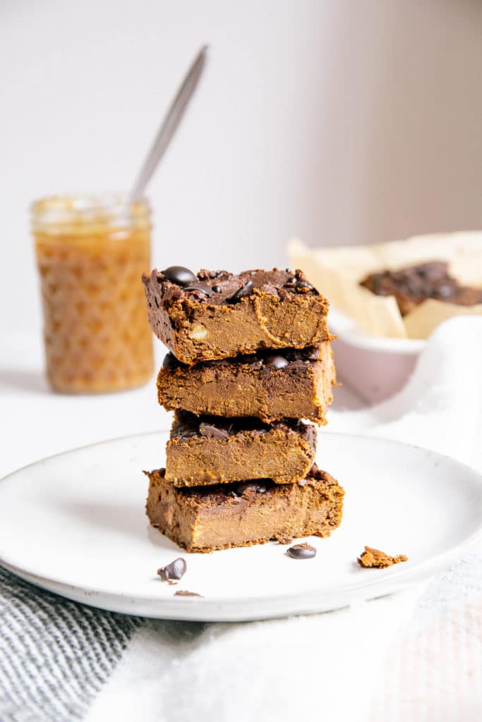 Ein Stapel vegane und zuckerfreie Protein-Brownies auf einem hellgrauen Teller mit einem Glas Dattelcreme und der Brownies-Backform im Hintergrund.