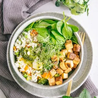 Gemüsereis mit gebratenem Tofu in einer graublauen Schüssel auf dazu passendem Teller mit einer goldenen Gabel darauf. Graues Küchentuch als Untergrund.