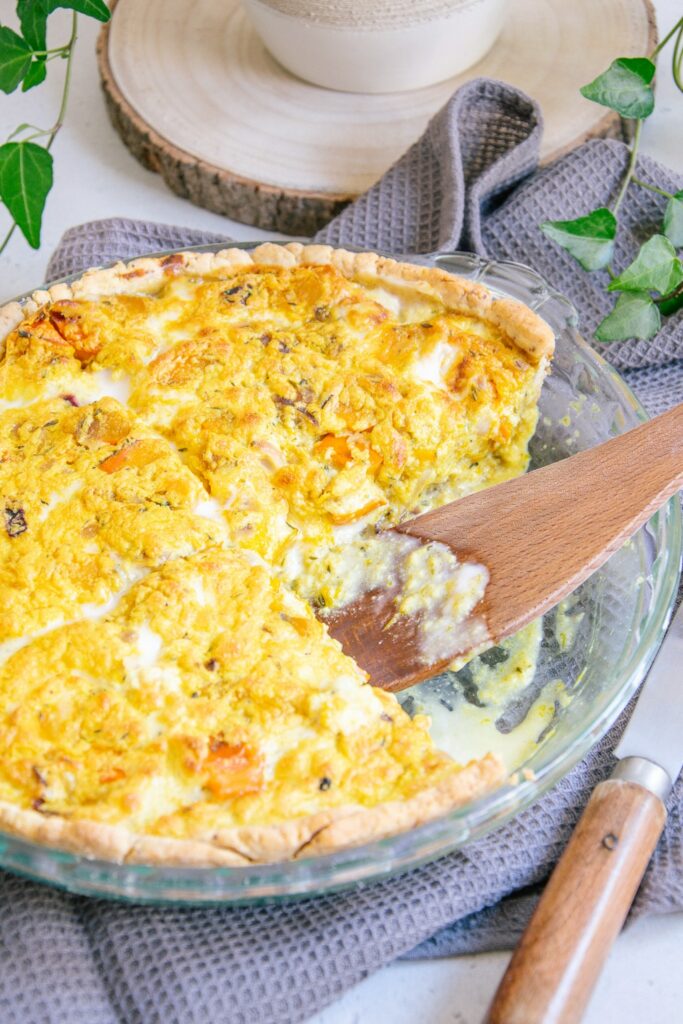 Vegane Kürbis-Quiche mit cremiger Füllung und hölzernem Küchenhelfer, der gerade ein Stück aus der Form genommen hat.