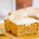 Ein Stück veganer zuckerfreier Kürbiskuchen fotografiert auf Augenhöhe, sodass die Vorderseite und eine Seite sichtbar sind. Detailaufnahme in orangen Herbstfarben.