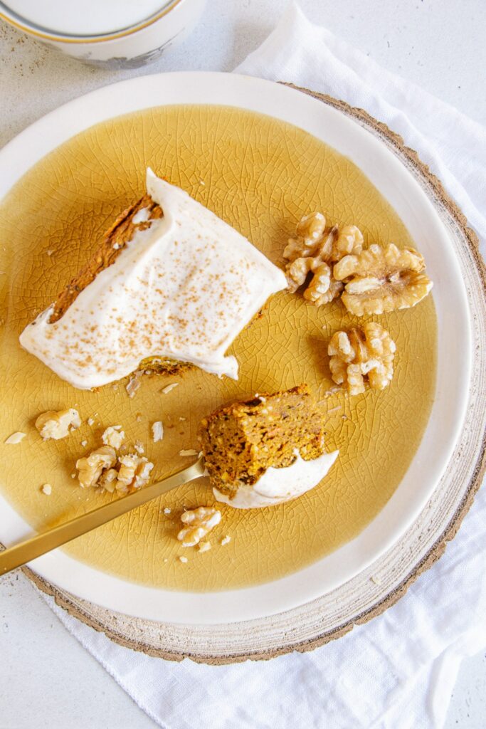 Ein Stück veganer zuckerfreier Kürbiskuchen auf einem dunkelorangen Teller, dekoriert mit Walnüssen. Eine goldene Gabel mit einem abgestochenen Stück Kuchen liegt auf dem Teller.