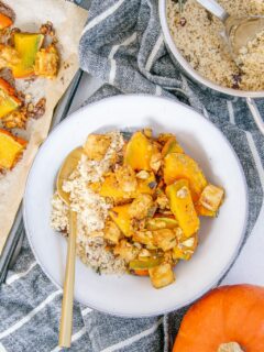 Ofen-Kürbis mit Knusper-Tofu und Couscous in einer graublauen Schüssel mit goldenem Löffel auf einem dunkelgrau-weiß gestreiften Küchentuch. Topf mit Couscous, Blech mit gebackenem Kürbis und ganzer Kürbis als Dekoration.