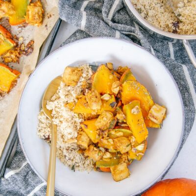 Ofen-Kürbis mit Knusper-Tofu und Couscous in einer graublauen Schüssel mit goldenem Löffel auf einem dunkelgrau-weiß gestreiften Küchentuch. Topf mit Couscous, Blech mit gebackenem Kürbis und ganzer Kürbis als Dekoration.