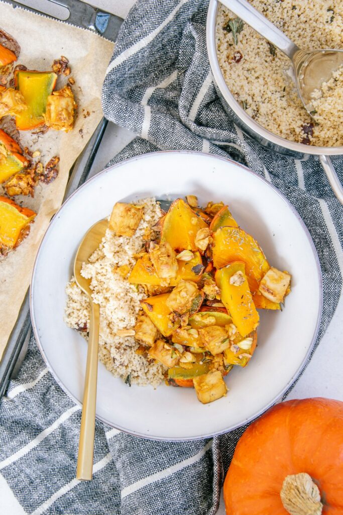 Ofen-Kürbis mit Knusper-Tofu und Couscous in einer graublauen Schüssel mit goldenem Löffel auf einem dunkelgrau-weiß gestreiften Küchentuch. Topf mit Couscous, Blech mit gebackenem Kürbis und ganzer Kürbis als Dekoration.