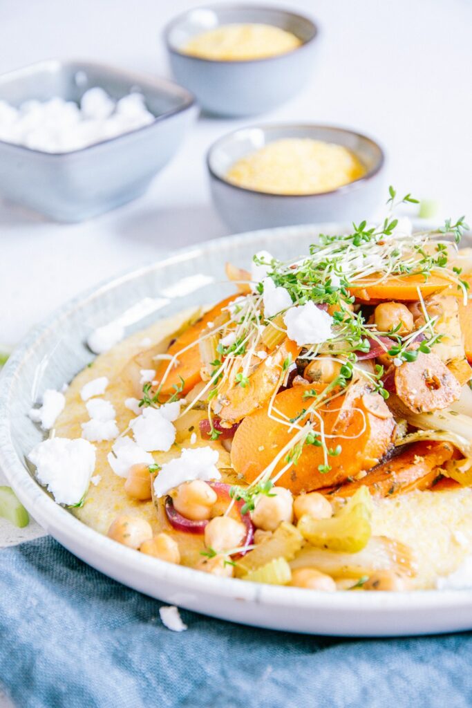 Nahaufnahme von Schmorgemüse mit Cremepolenta im 45-Grad-Winkel. Garniert mit veganem Feta und Kresse.