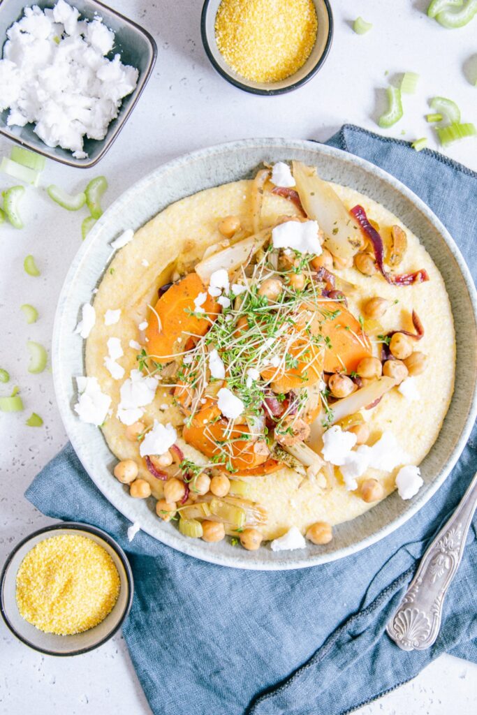 Schmorgemüse mit Cremepolenta auf einem grau-grünen Teller aus der Vogelperspektive fotografiert. Blaues Leinentuch und heller Untergrund. Kleine Schälchen mit Polenta und veganem Feta als Dekoration.