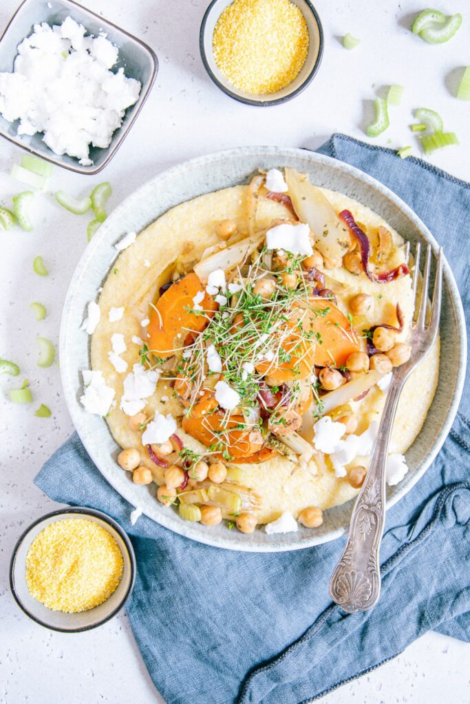 Schmorgemüse mit Cremepolenta, Kichererbsen, veganem Feta und Kresse auf einem grau-grünen Teller mit einer rustikalen Gabel aus der Vogelperspektive fotografiert.