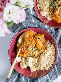 Schnelles Pfannengemüse mit würziger Sauce, Couscous und Tempeh in einem tiefen dunkelroten Teller mit goldenem Löffel. Aufnahme aus der Vogelperspektive.