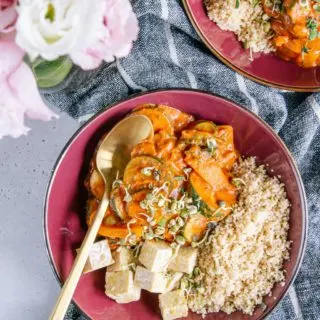 Schnelles Pfannengemüse mit würziger Sauce, Couscous und Tempeh in einem tiefen dunkelroten Teller mit goldenem Löffel. Aufnahme aus der Vogelperspektive.