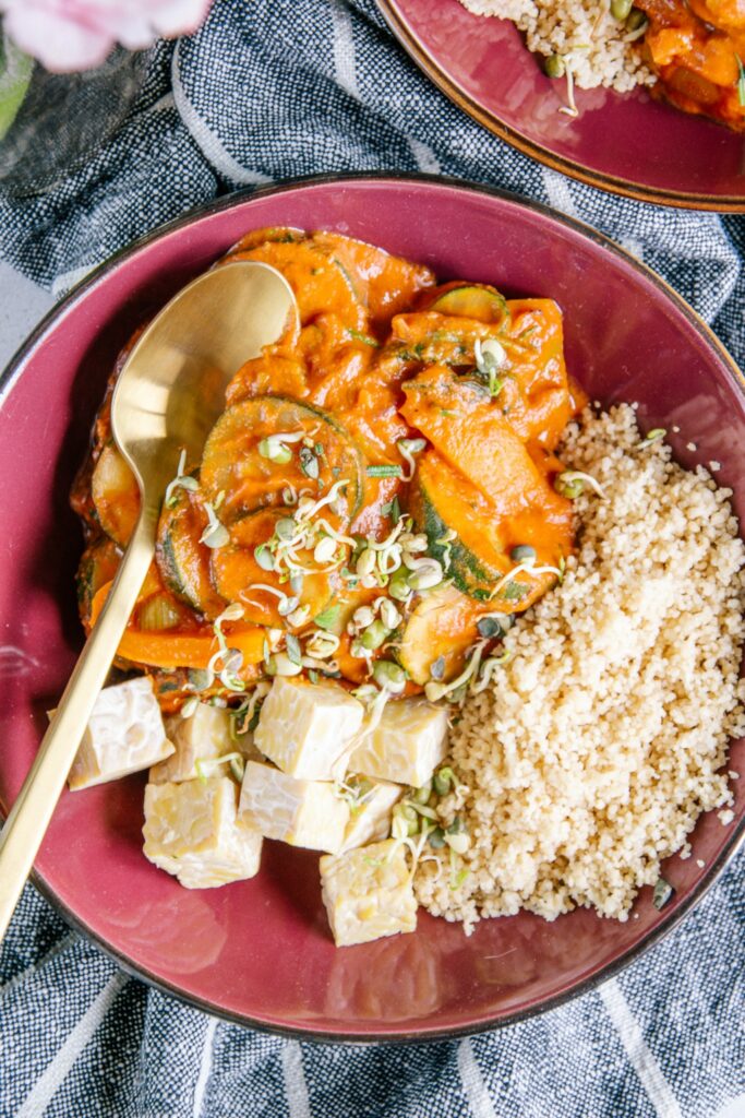 Pfannengemüse mit würziger Sauce, Couscous und Tempeh zu je einem Drittel auf einem tiefen dunkelroten Teller aufgeteilt. Goldener Löffel im Pfannengemüse. Aufnahme aus der Vogelperspektive.