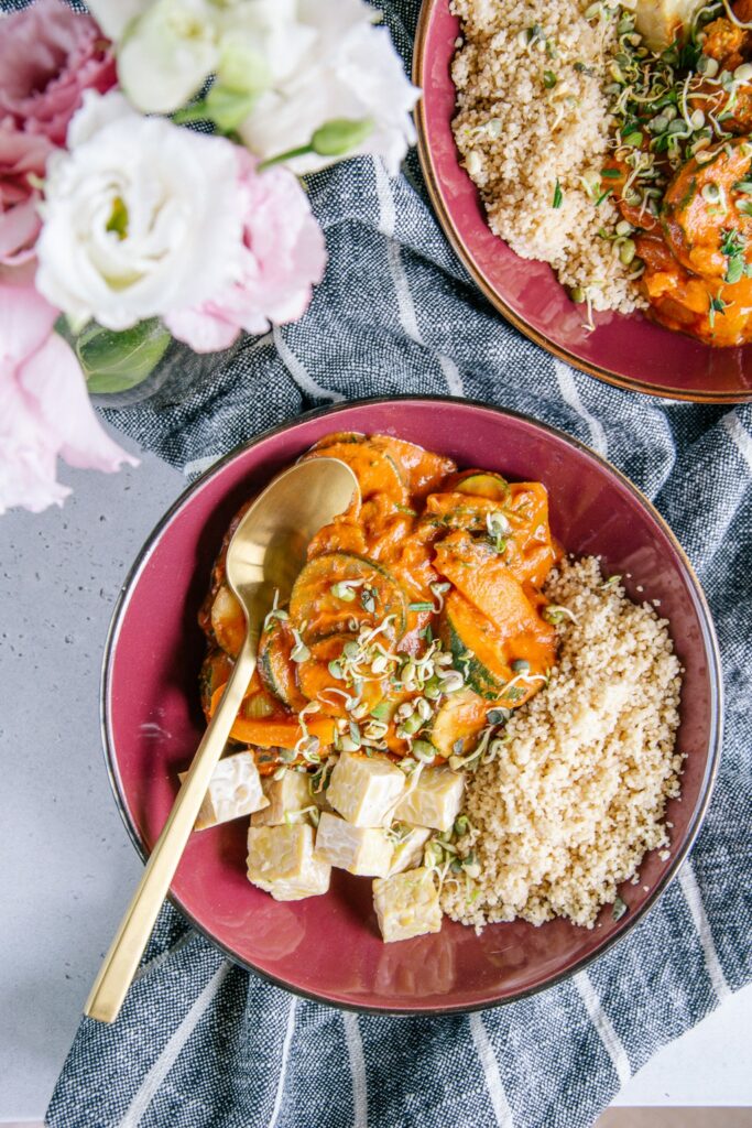 Schnelles Pfannengemüse mit würziger Sauce, Couscous und Tempeh in einem tiefen dunkelroten Teller mit goldenem Löffel. Aufnahme aus der Vogelperspektive.