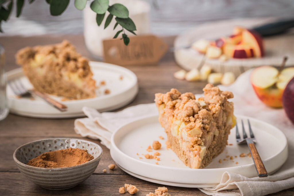 Zwei Teller mit Streuselkuchen und ein Schneidebrett mit einem gewürfelten Apfel im Hintergrund