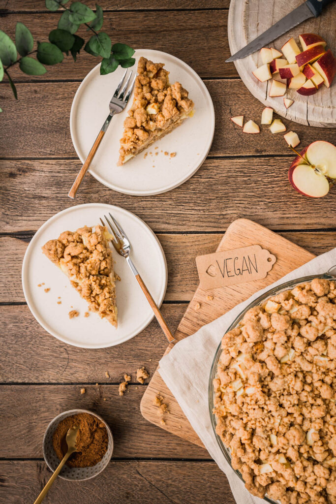 Zwei Stücke Apfel Streuselkuchen auf cremeweißen Tellern, daneben Kuchengabeln mit Holzgriff