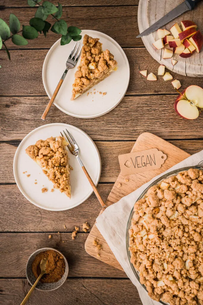 Zwei Stücke Apfel Streuselkuchen auf cremeweißen Tellern, daneben Kuchengabeln mit Holzgriff