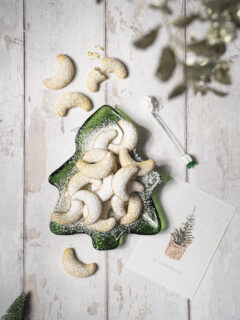 Mit Staubzucker bestreute Vanillekipferl auf einem gläsernen grünen Keksteller in Tannenbaum-Form.