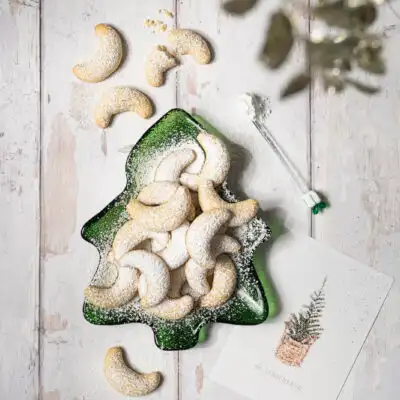 Mit Staubzucker bestreute Vanillekipferl auf einem gläsernen grünen Keksteller in Tannenbaum-Form.