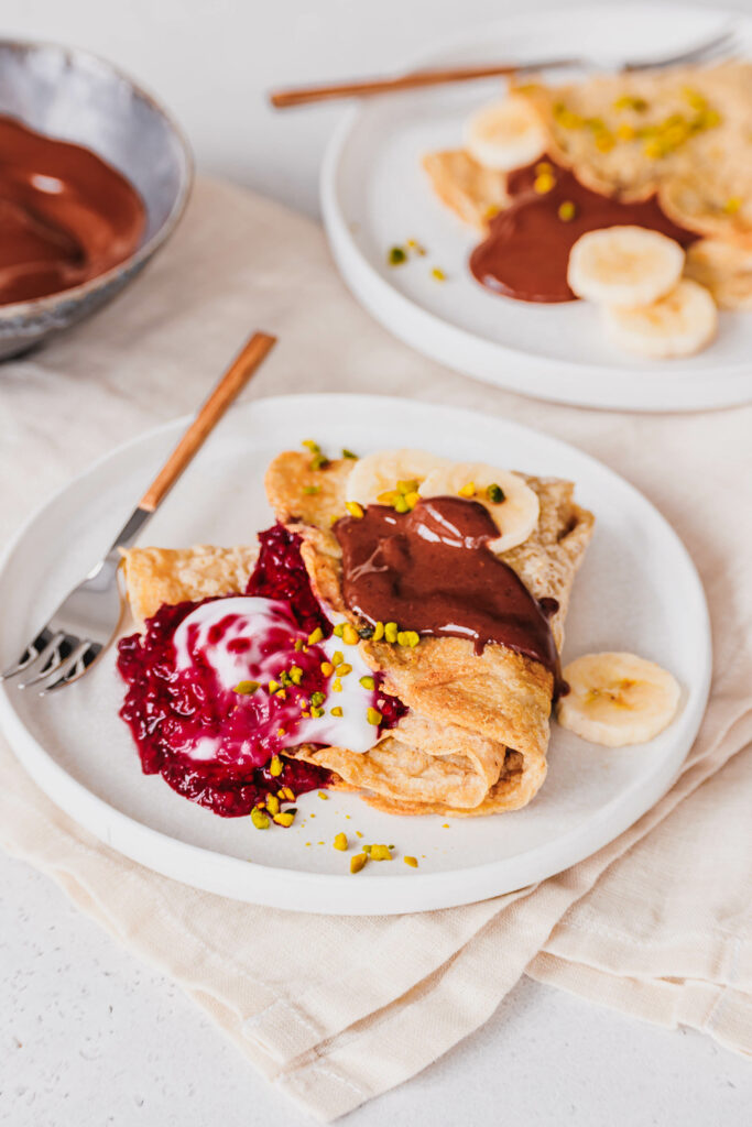 Haferflocken-Pfannkuchen mit Himbeer-Chia-Marmelade, Kokosjoghurt, Schoko-Mandelcreme, Bananenscheiben und gehackten Pistazien