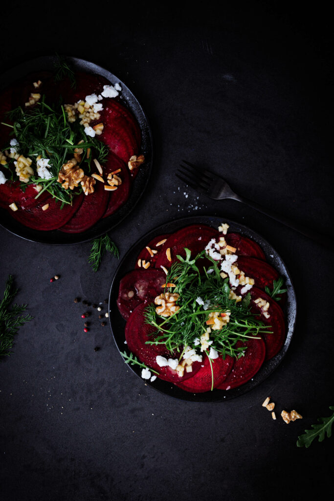 Rohe Rote-Rüben-Scheiben mit Rucola, Apfel-Dressing, veganem Feta und Nüssen auf zwei schwarzen Tellern.